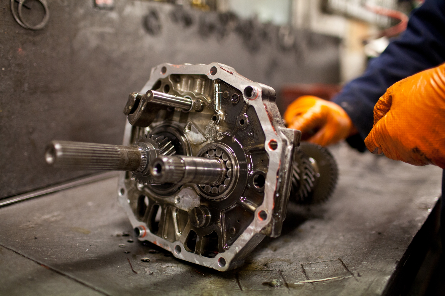 Mechanic wearing orange gloves working on a disassembled car transmission placed on a workshop table, showing exposed gears and metal housing.