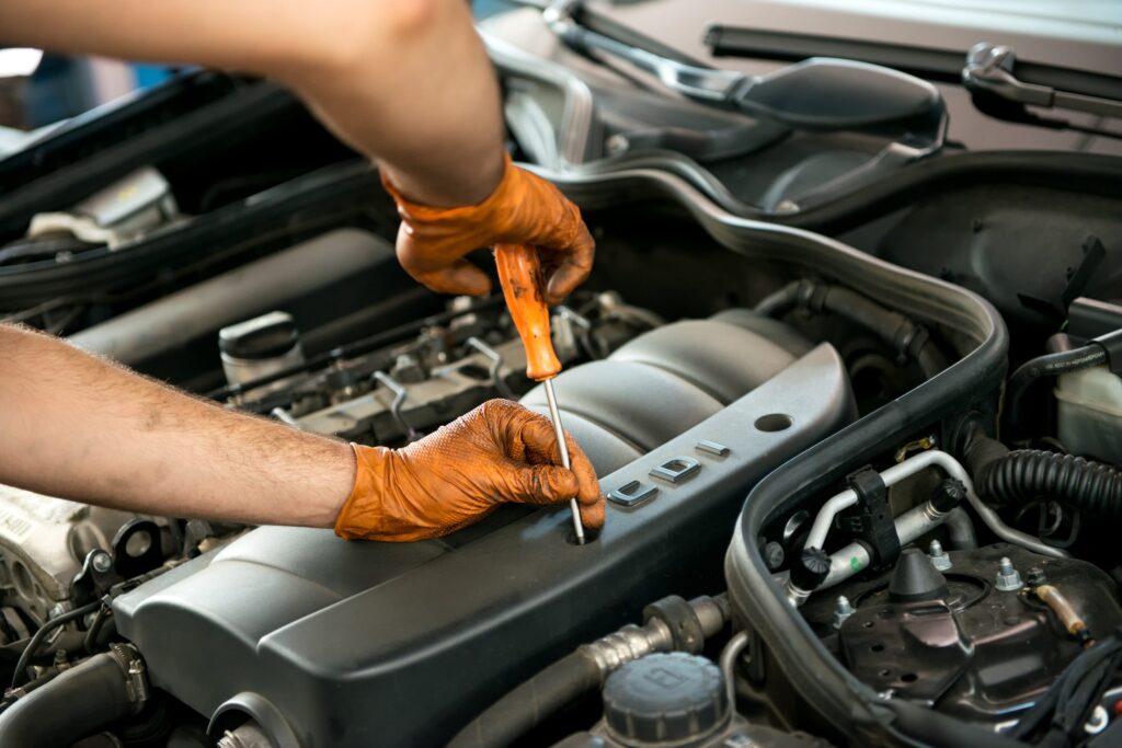 Mechanic wearing orange gloves using a screwdriver to work on a car engine, performing maintenance or repair tasks.