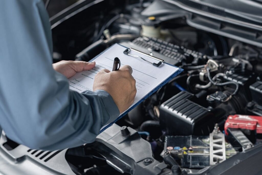 Mechanic inspecting car engine with a checklist on a clipboard, performing vehicle maintenance or service evaluation.