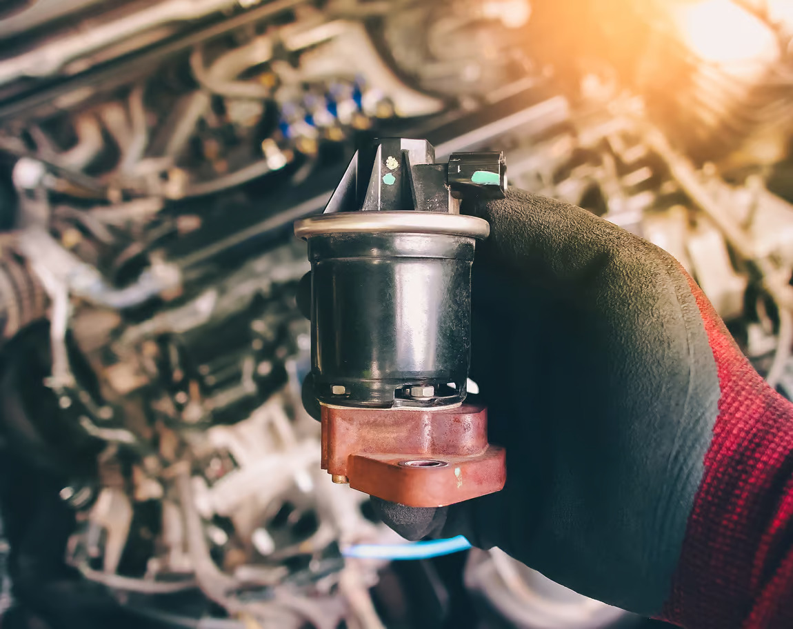 Mechanic wearing a glove holding a car engine sensor or valve component in front of an open engine bay.